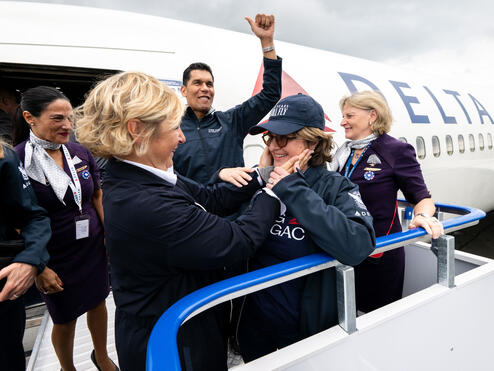 Delta employees and volunteers stand on an airport boarding ramp beside a Delta aircraft, welcoming a passenger as they exit the plane.
