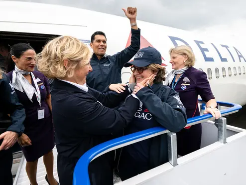 Delta employees and volunteers stand on an airport boarding ramp beside a Delta aircraft, welcoming a passenger as they exit the plane.
