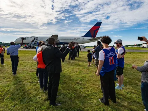 Spectators stand on grass watching a Delta airplane taxi on the runway at an airport.