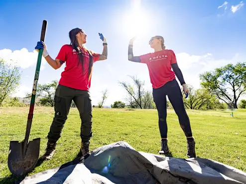 Two volunteers in red “Delta in the Community” shirts high-five while working outdoors, one holding a shovel on a sunny grassy field with trees in the background.