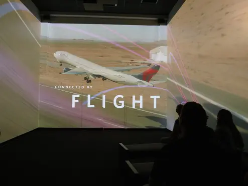Silhouetted audience in a dark room viewing a multi-wall projection of a plane taking off, with the text “CONNECTED BY FLIGHT” overlaid.