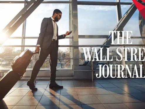 Traveler in a suit walks through a sunlit airport terminal, pulling a rolling suitcase and looking at a phone, with “The Wall Street Journal” text overlaid.