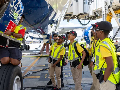 Students in safety vests gather beneath a Delta aircraft as an aviation professional points out features near the landing gear on the airport ramp.