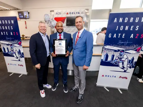 Three people pose at a Delta airport gate ceremony, with the center person holding a commemorative plaque. Banners on both sides read “Andruw Jones Gate A25,” featuring baseball-themed artwork and Delta branding.