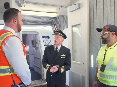 Three airport employees standing and talking near an aircraft boarding door, including a pilot in uniform and two workers wearing high-visibility safety vests.