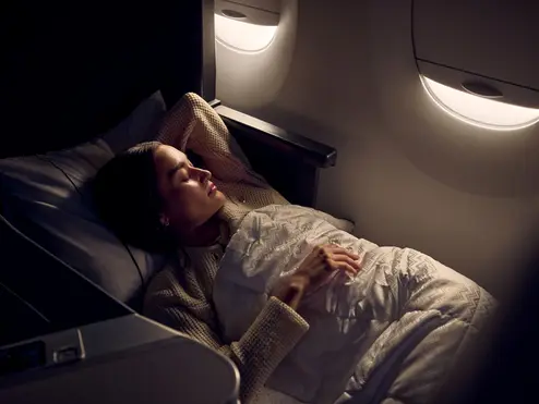  Passenger resting in a lie-flat Delta One seat with bedding, illuminated by soft cabin lighting near an aircraft window.