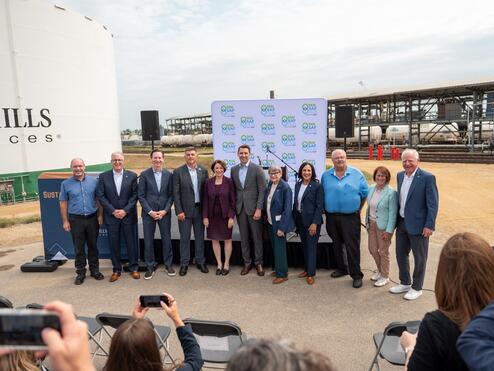 Group photo of event attendees standing in front of an industrial biofuels facility, with storage tanks, railcars, and a branded backdrop visible behind them while audience members take photos in the foreground.