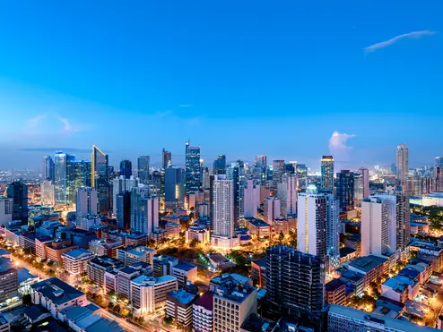 A panoramic view of a modern city skyline at dusk, with illuminated high-rise buildings, busy streets, and a bright blue evening sky.