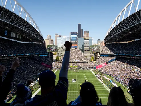 Fans in a packed football stadium watch the game from the stands, with the field and Seattle skyline visible beyond the open end of the venue.