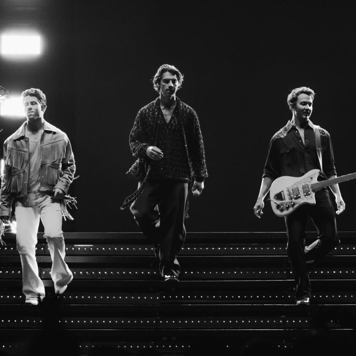 The Jonas Brothers walking on stage stairs during concert in black and white.