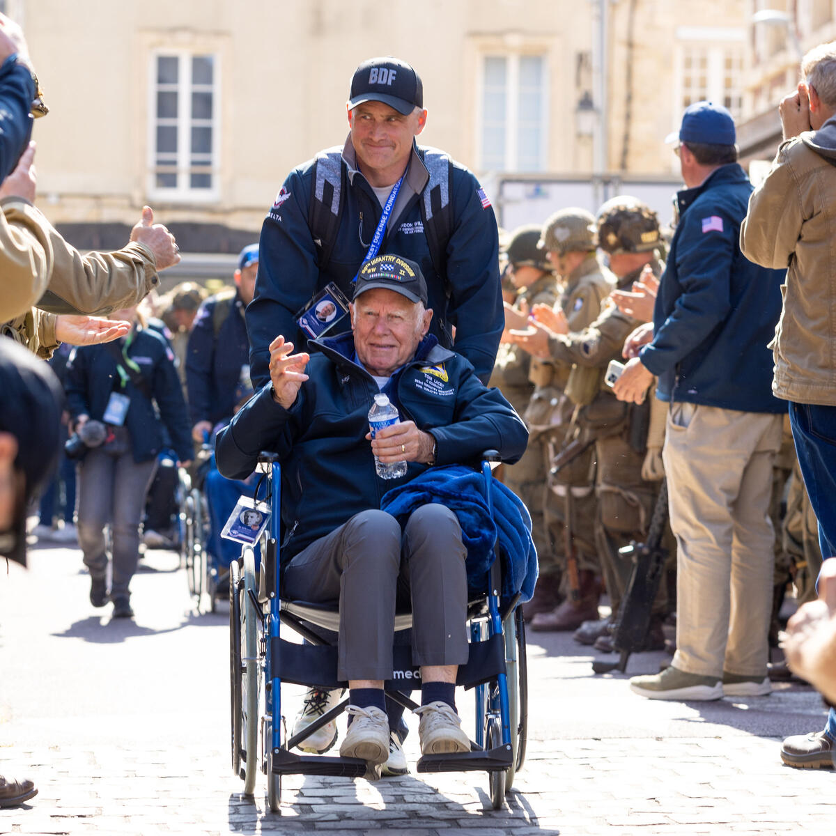 Army veteran Tom Lacey and his caretaker, Air Force veteran and Atlanta-based 717 Capt. Brandon Williams, wave to fans at a parade in Carentan.