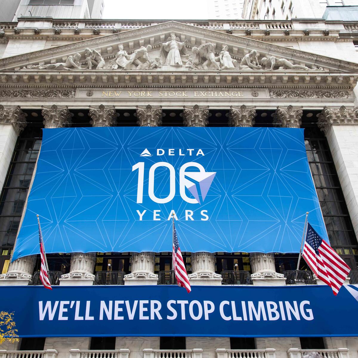 Exterior shot of NYSE building with a large, rectangular banner displaying Delta's centennial logo and a smaller banner below that reads We'll Never Stop Climbing