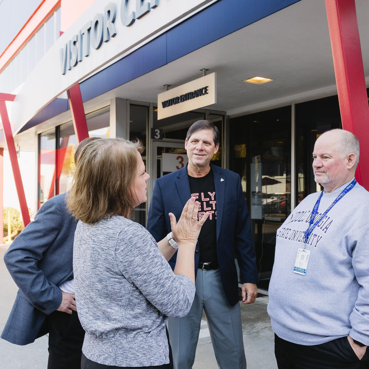 Four people talking outside a Visitor Center entrance with red beams; one wears a ‘Fly Delta Jets’ shirt, another a Middle State sweatshirt.
