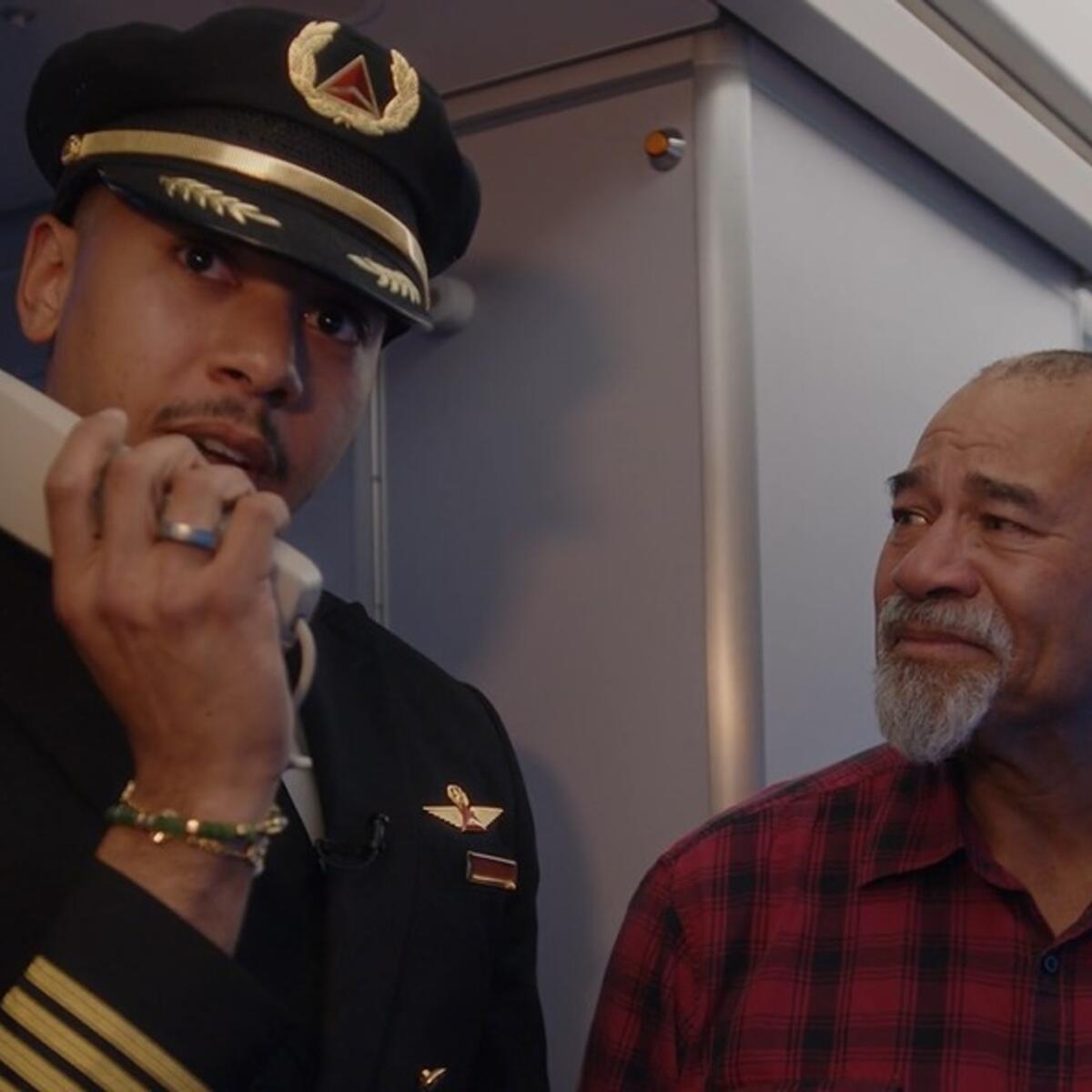Airline pilot in uniform stands inside an aircraft cabin near the galley, holding an intercom handset. Another person wearing a red plaid shirt is standing beside the pilot.
