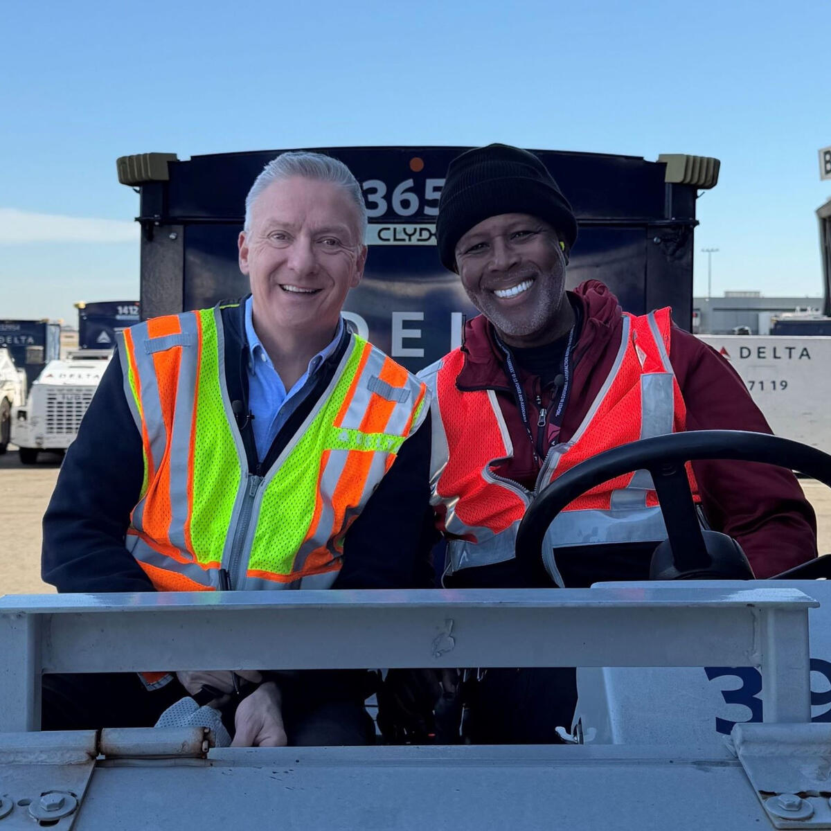 Two workers in high-visibility vests sitting on an airport ground vehicle near Delta equipment and a boarding gate.