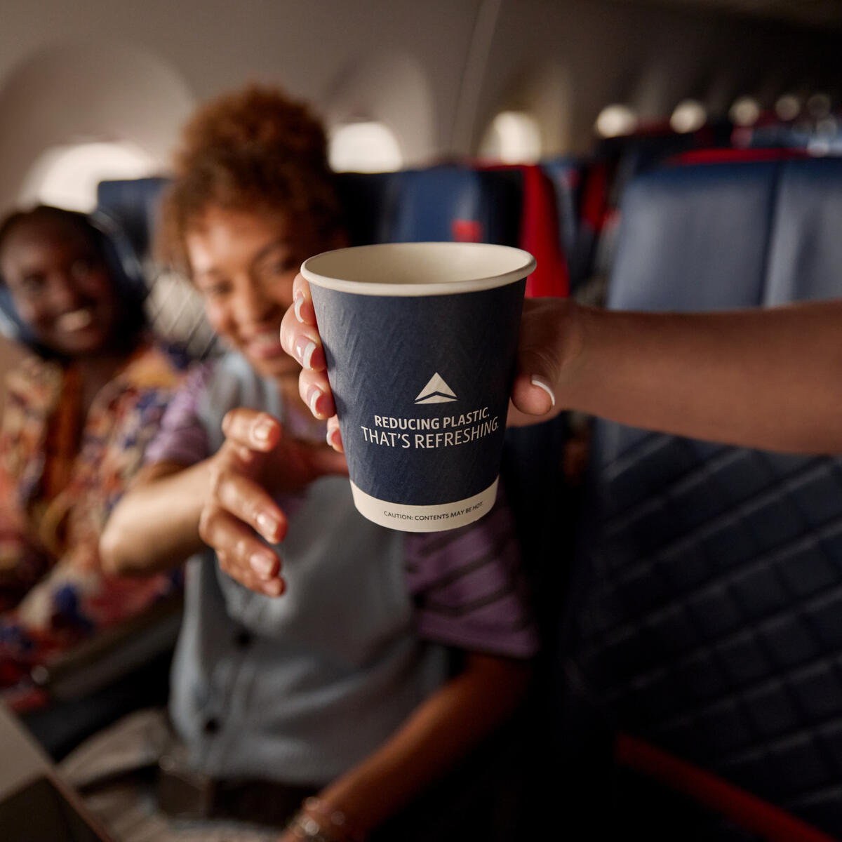 Delta's new paper cup in flight attendant's hand, ready to be handed to customer who is seated on board an aircraft and reaching for the cup.