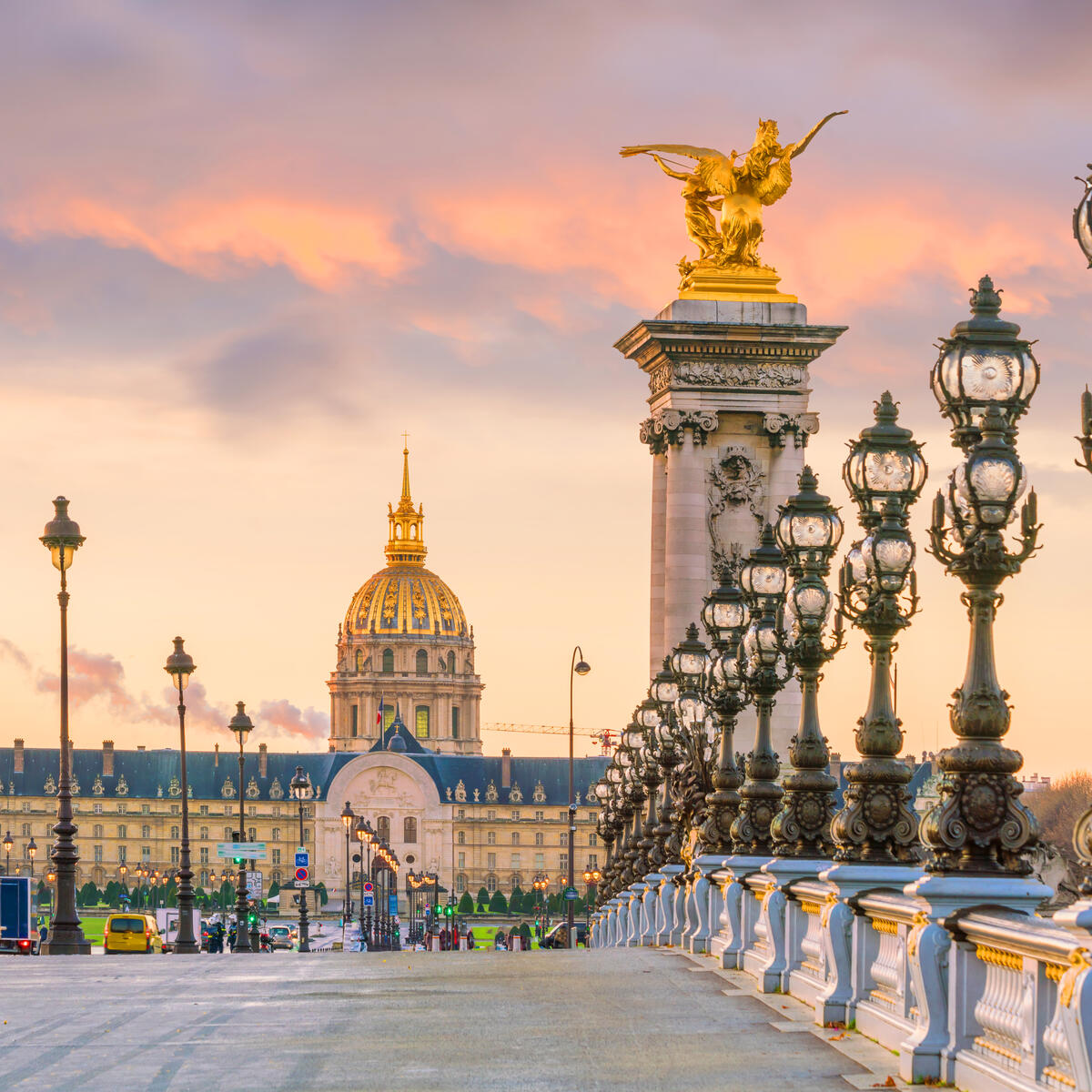 Ornate bridge lined with lamps at sunset, facing a domed historic building in the distance.