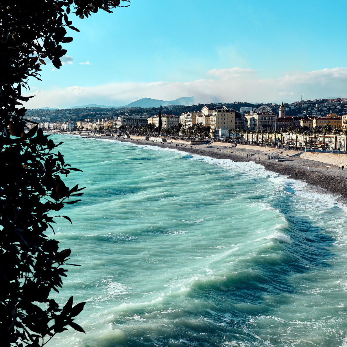 Waves rolling toward a long beachfront lined with buildings under a bright blue sky.