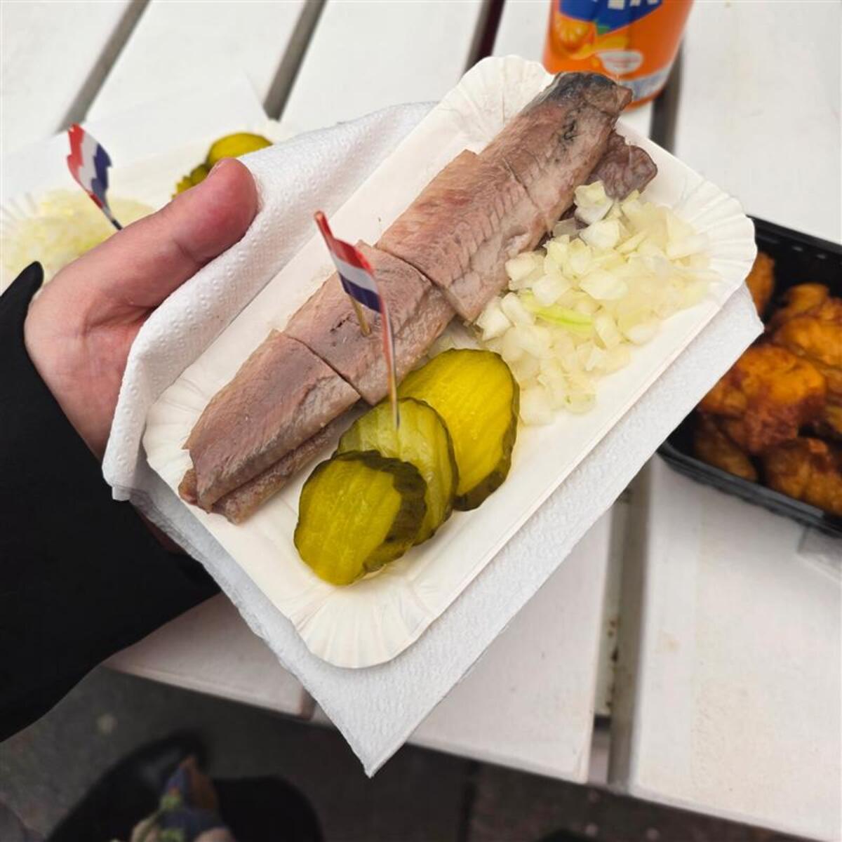  tray of Dutch-style raw herring served with chopped onions and pickle slices, held in one hand with a small Dutch flag toothpick.