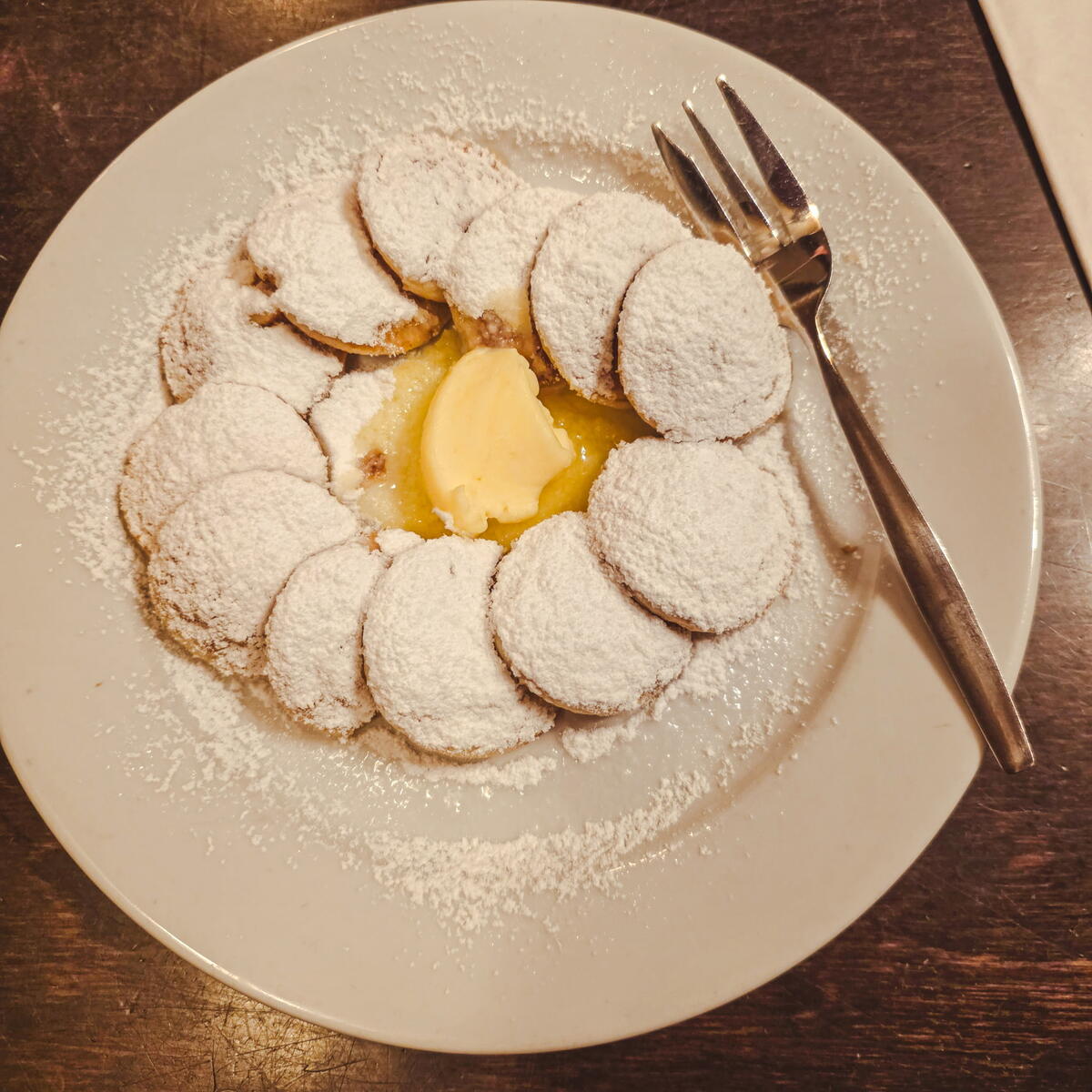 A plate of small, round pancakes dusted heavily with powdered sugar, served with a pat of butter in the center and a fork on the side.