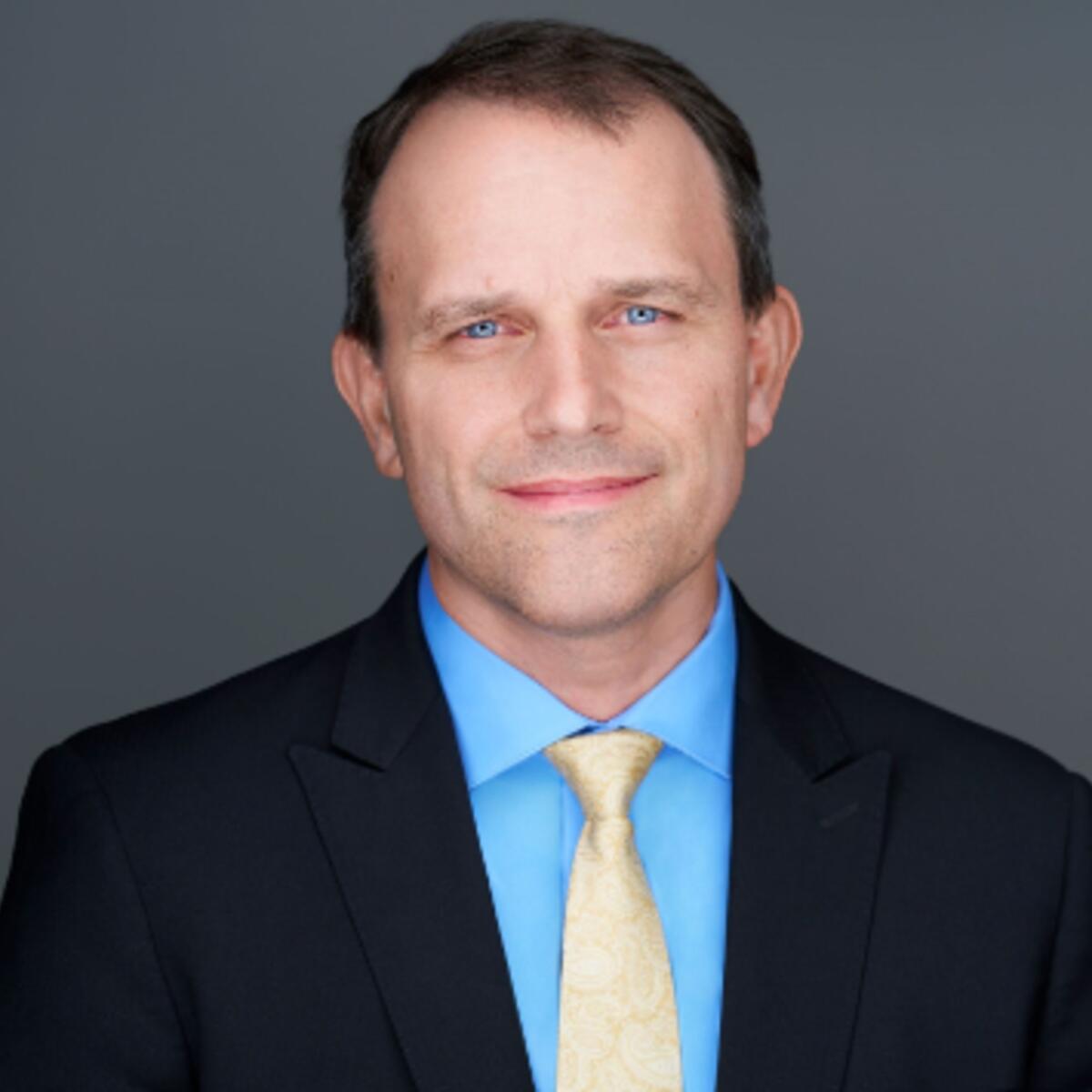 Headshot of Matt Schrag, wearing a dark suit jacket, light blue dress shirt and light yellow tie, posed against a plain gray background.