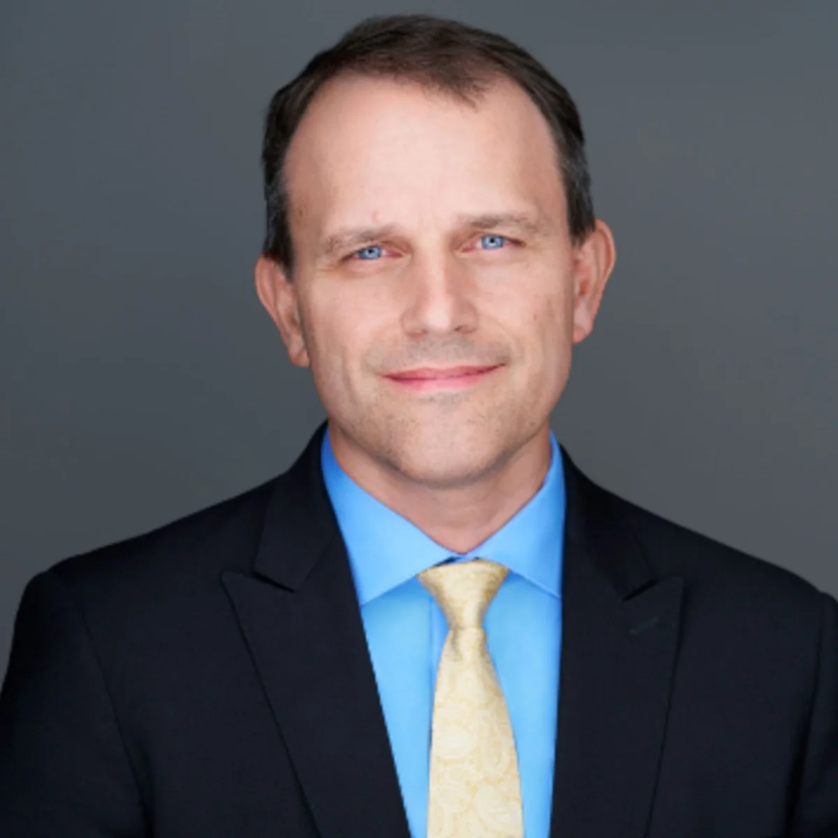 Headshot of Matt Schrag, wearing a dark suit jacket, light blue dress shirt and light yellow tie, posed against a plain gray background.