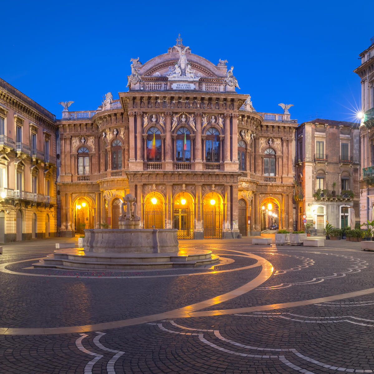 Illuminated historic opera house facing an empty patterned square at dusk.