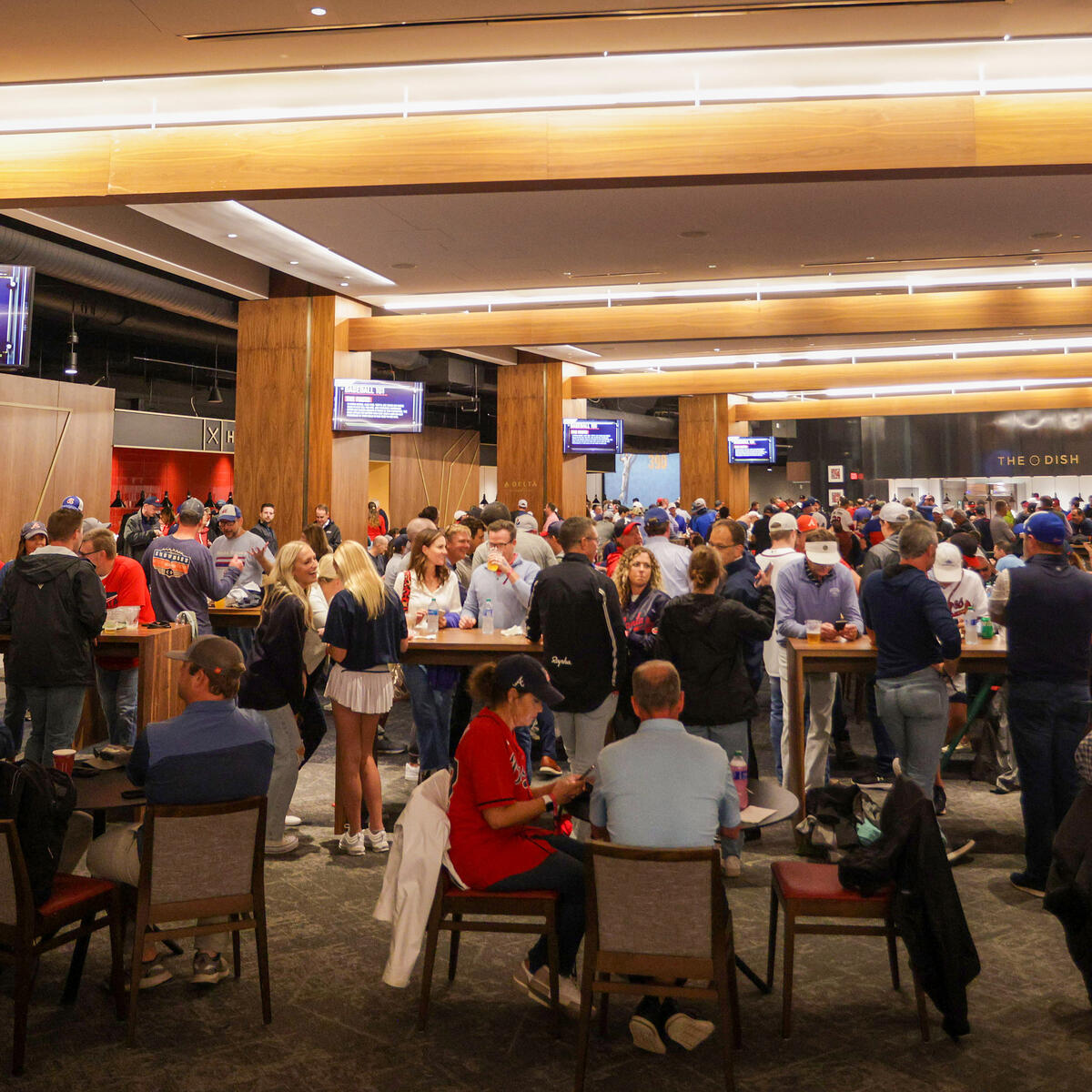 A crowded indoor stadium lounge with fans standing and sitting at tables beneath wood-paneled ceilings and multiple TV screens.