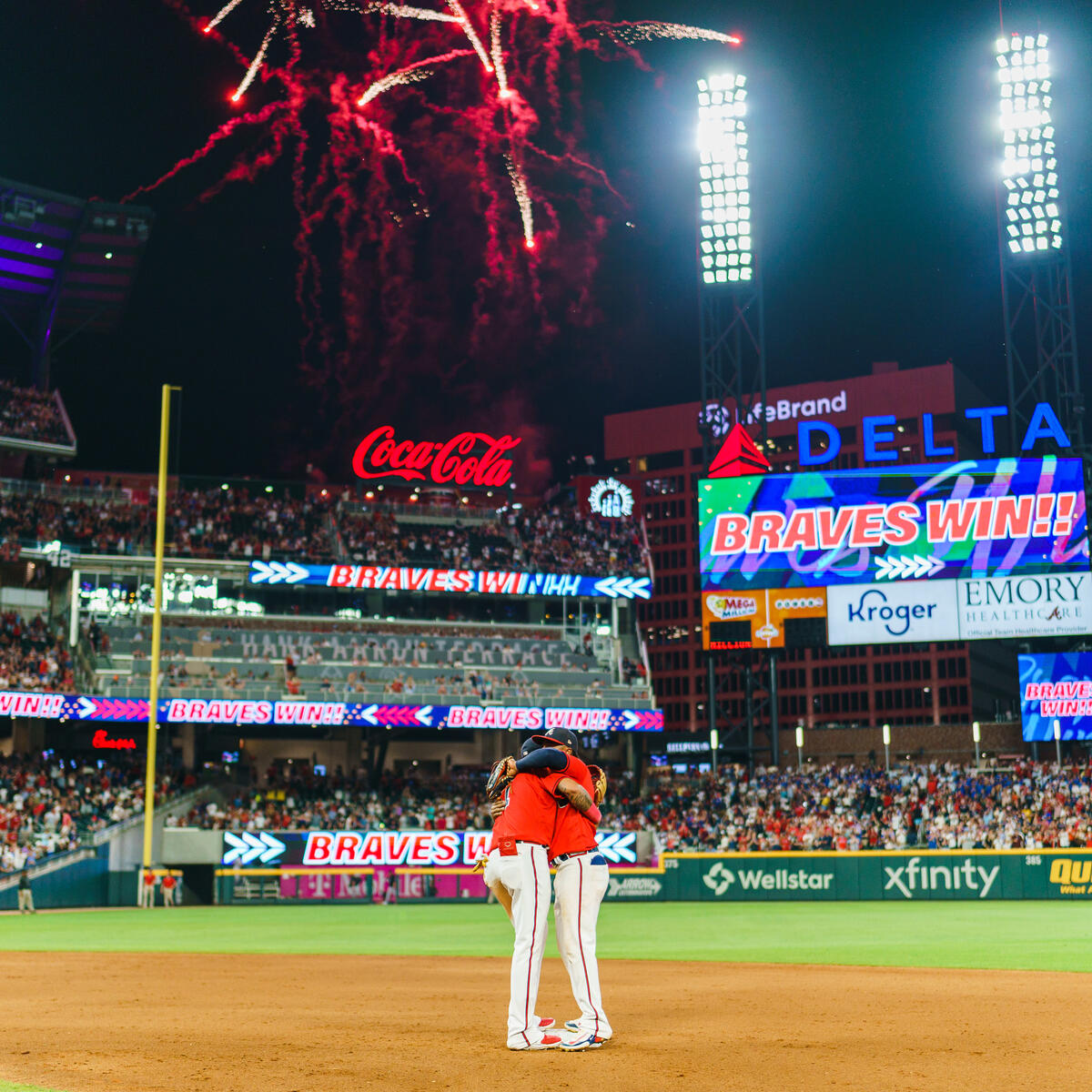 Fireworks light up a baseball stadium at night as two players stand together on the infield, with a scoreboard displaying “Braves Win” and a packed crowd in the background.