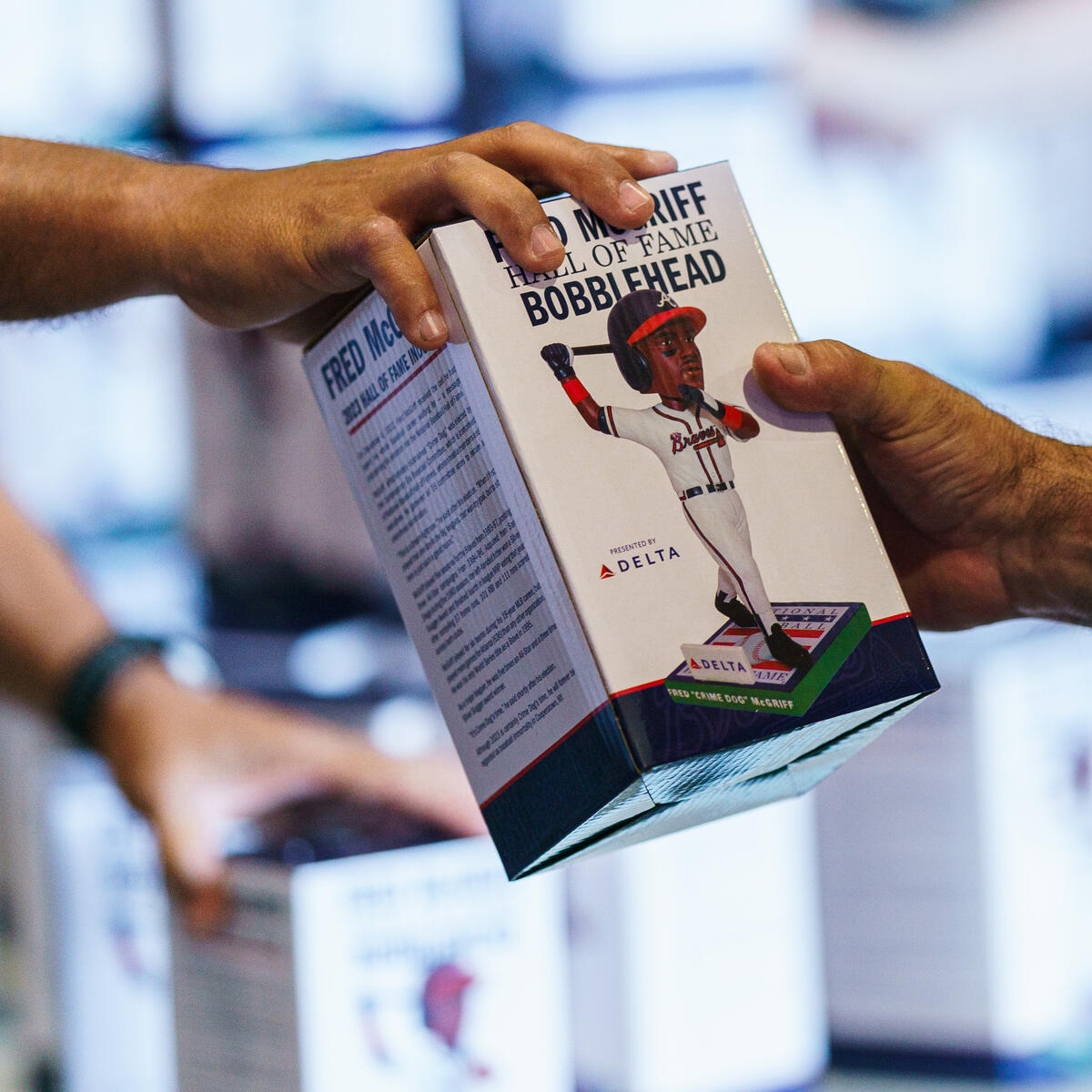 Two hands exchange a boxed “Hall of Fame Bobblehead,” showing a baseball player figurine on the packaging, with stacks of similar boxes in the background.