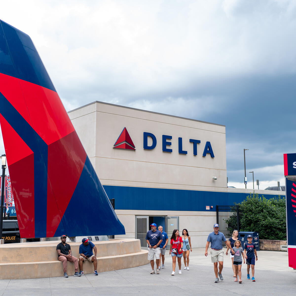 People walk outside a stadium concourse near a large Delta airplane tail sculpture and a Delta-branded building, with a Braves merchandise shop nearby.
