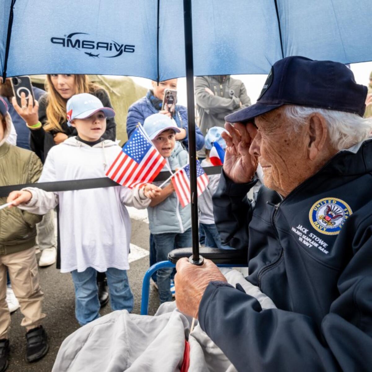 An older person in a veteran’s jacket sits under an umbrella while children hold small American flags nearby during an outdoor commemorative event.
