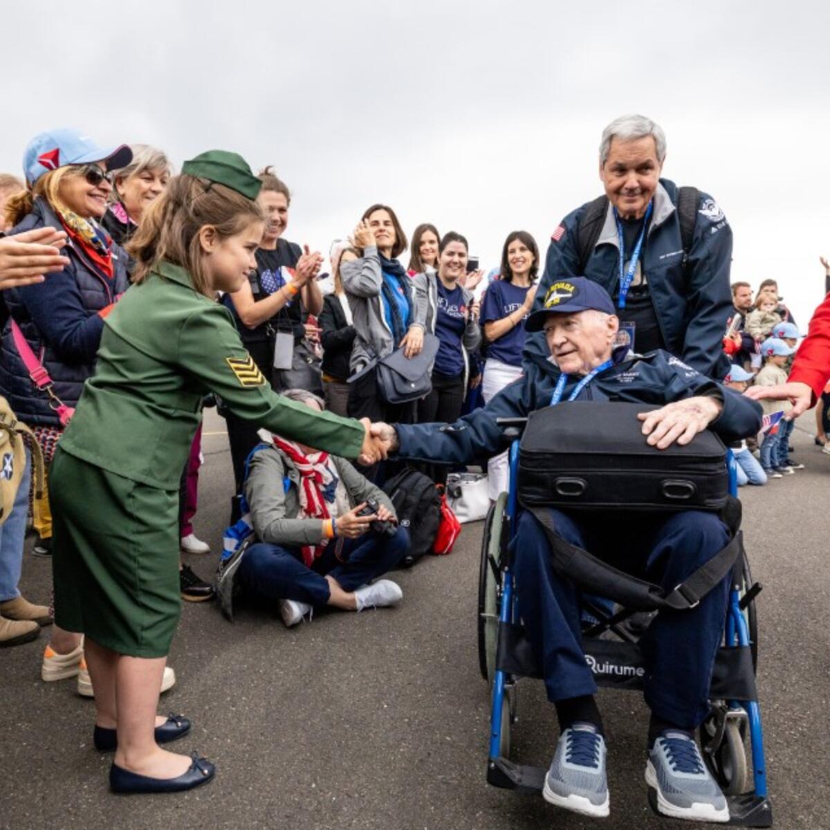 A seated veteran in a wheelchair shakes hands with a child in a green uniform as a crowd lines the roadway, applauding during a commemorative event.