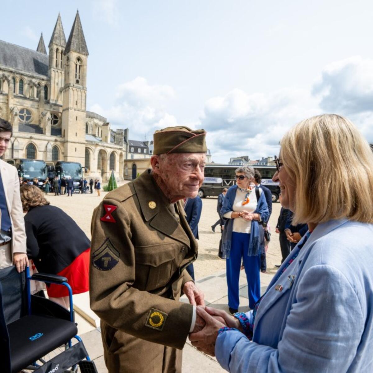A uniformed World War II–era veteran shakes hands with an attendee in front of a historic stone church, with a small group of people gathered nearby at an outdoor commemorative event.
