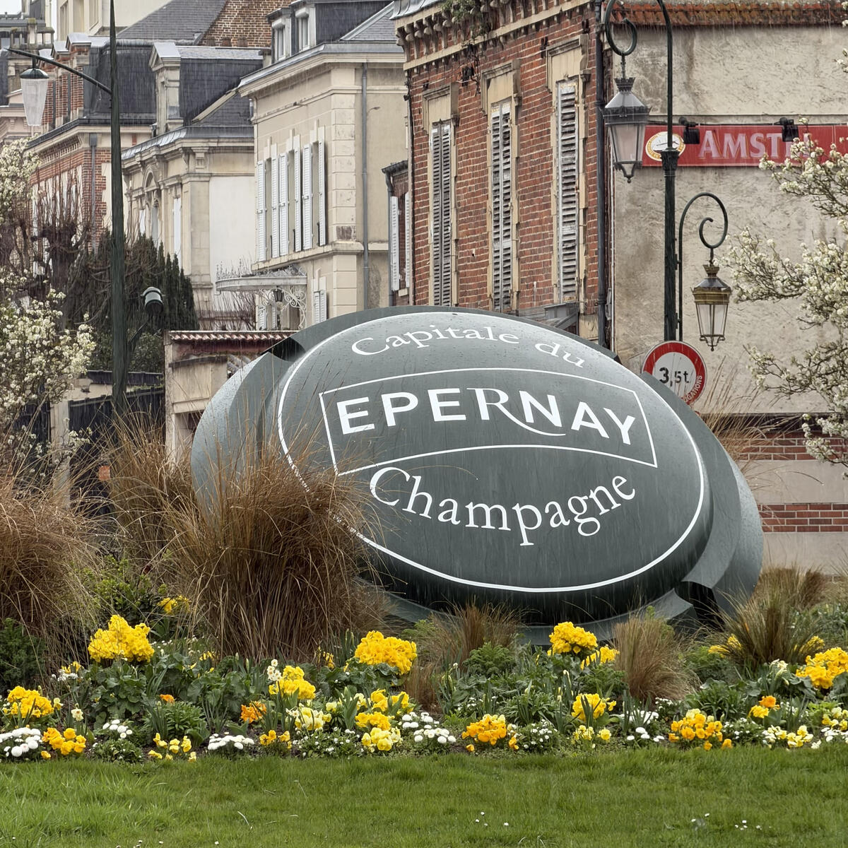 A large, green, shield-shaped monument with the white text "Capitale du Champagne, EPERNAY" sits in the middle of a floral roundabout, surrounded by yellow flowers and classic French buildings under a cloudy sky.