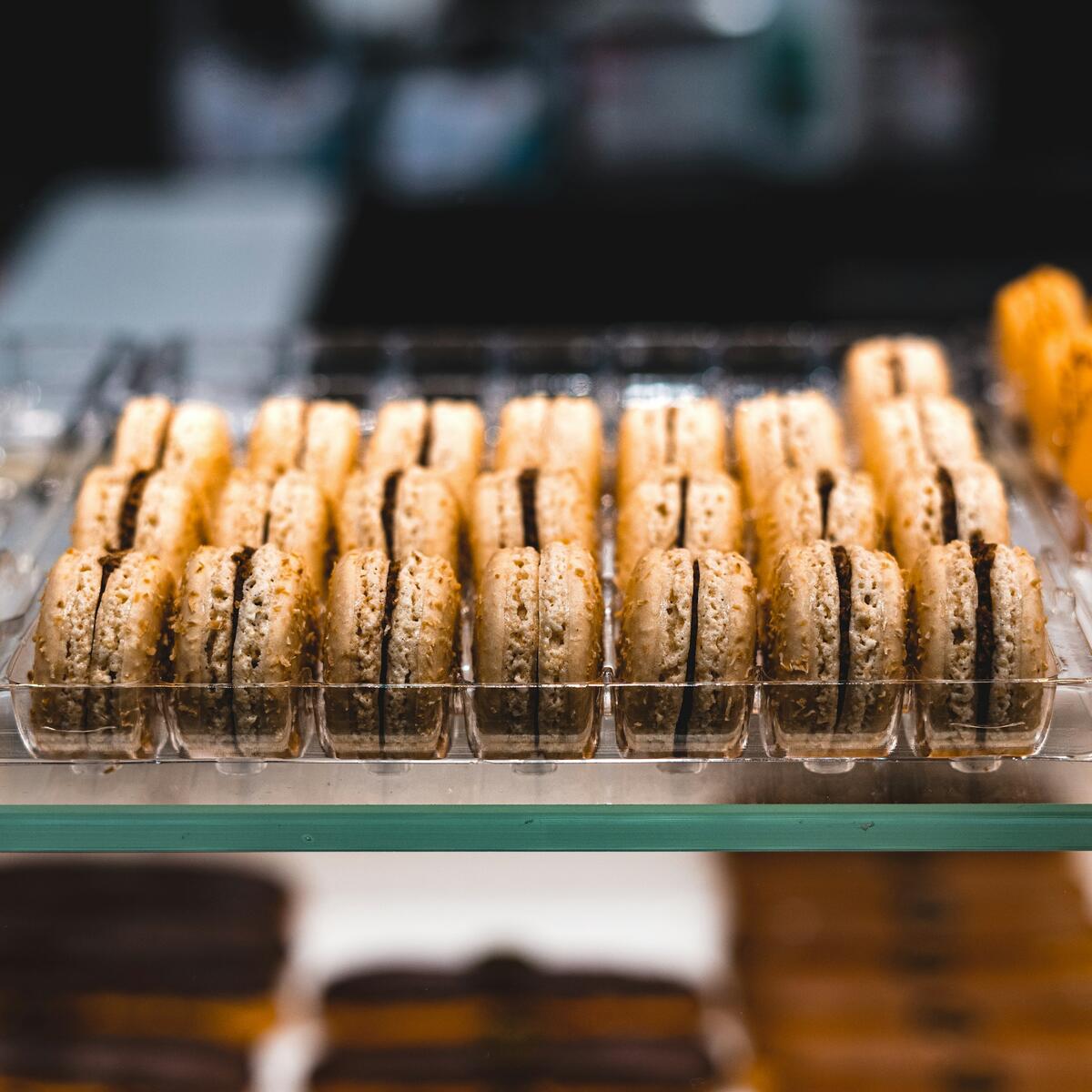A neatly arranged clear plastic tray filled with light-colored, sandwich-style macarons containing a dark brown filling, displayed in a bakery case.