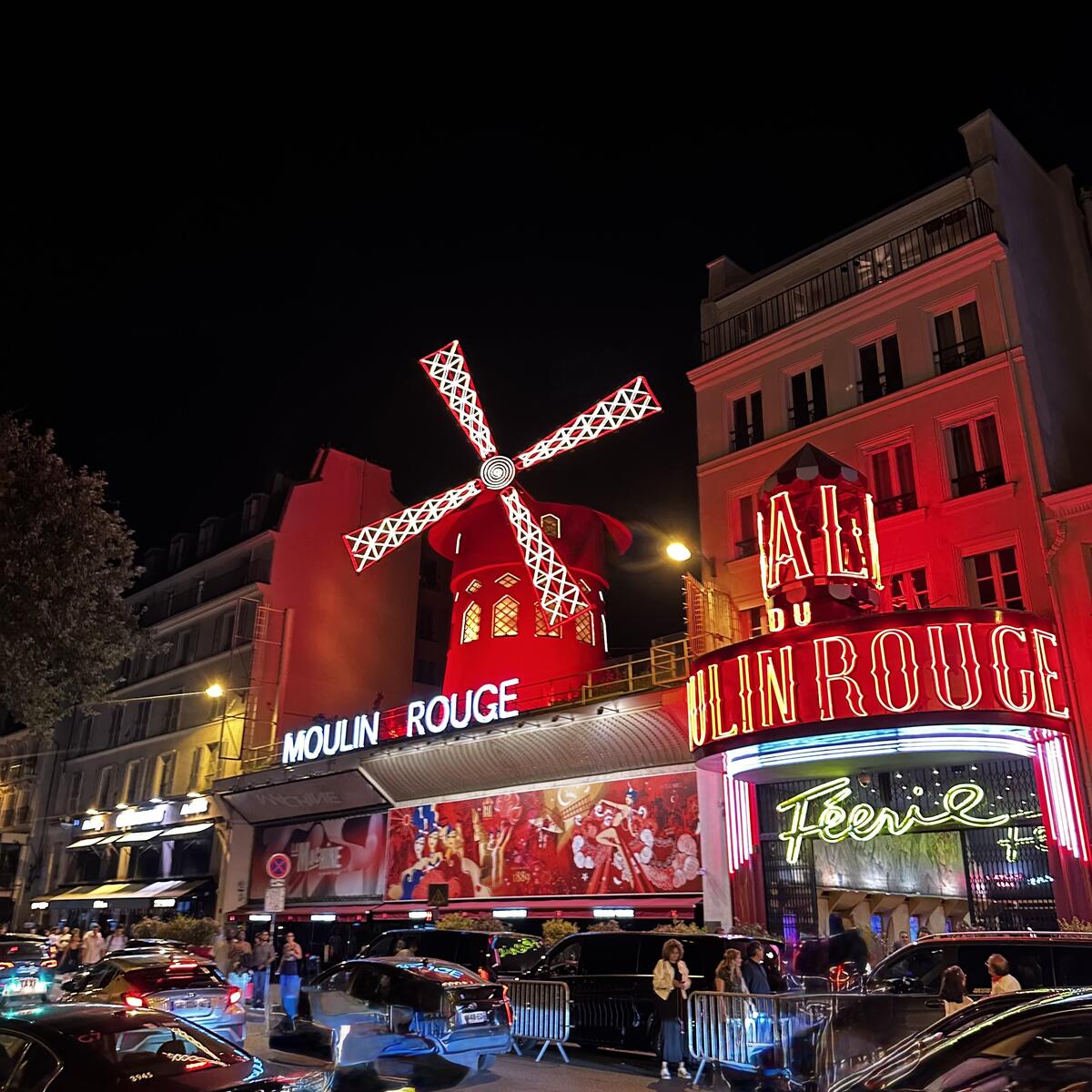 The iconic Moulin Rouge cabaret in Paris, with its illuminated red windmill and "FÉERIE" sign, bustling with people and traffic on a dark night.