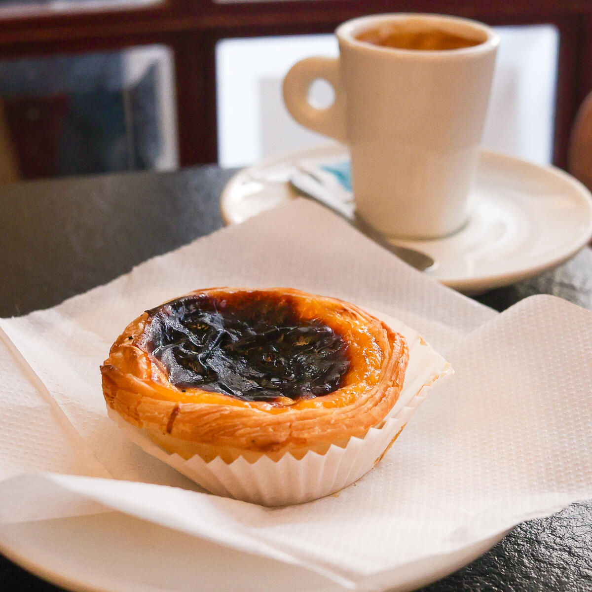 Portuguese custard tart (pastel de nata) on a napkin atop a café table, with a small cup of espresso on a saucer softly blurred in the background.