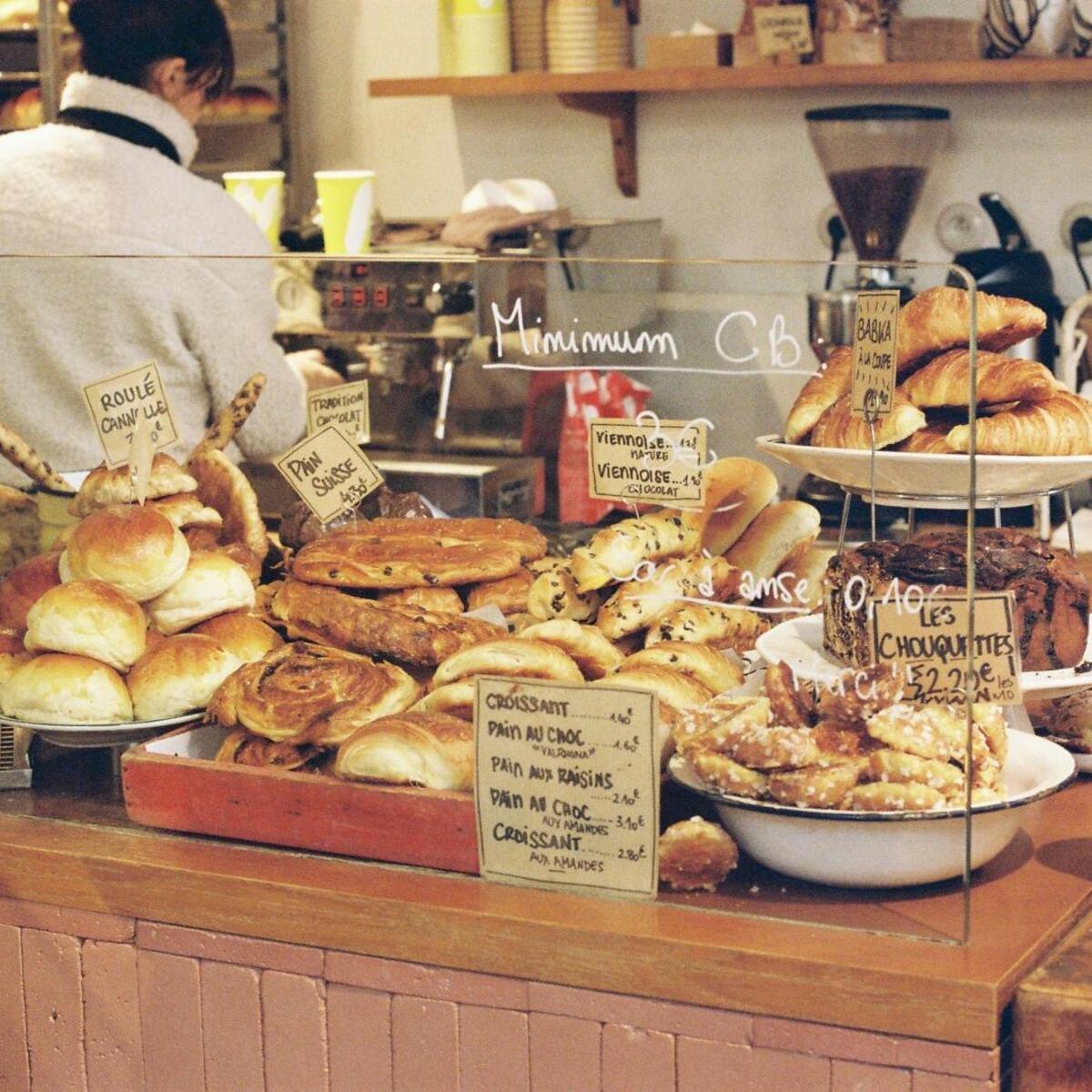 A well-stocked glass display case in a bakery, showcasing various golden French pastries like croissants, pain au chocolat, chouquettes, and large brioche, with handwritten price signs in French and "Minimum CB" text. A person is working in the background.