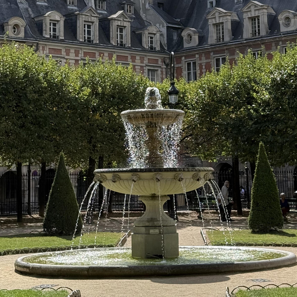 Stone fountain in a leafy Paris square, framed by manicured trees and historic red-brick buildings with slate roofs.