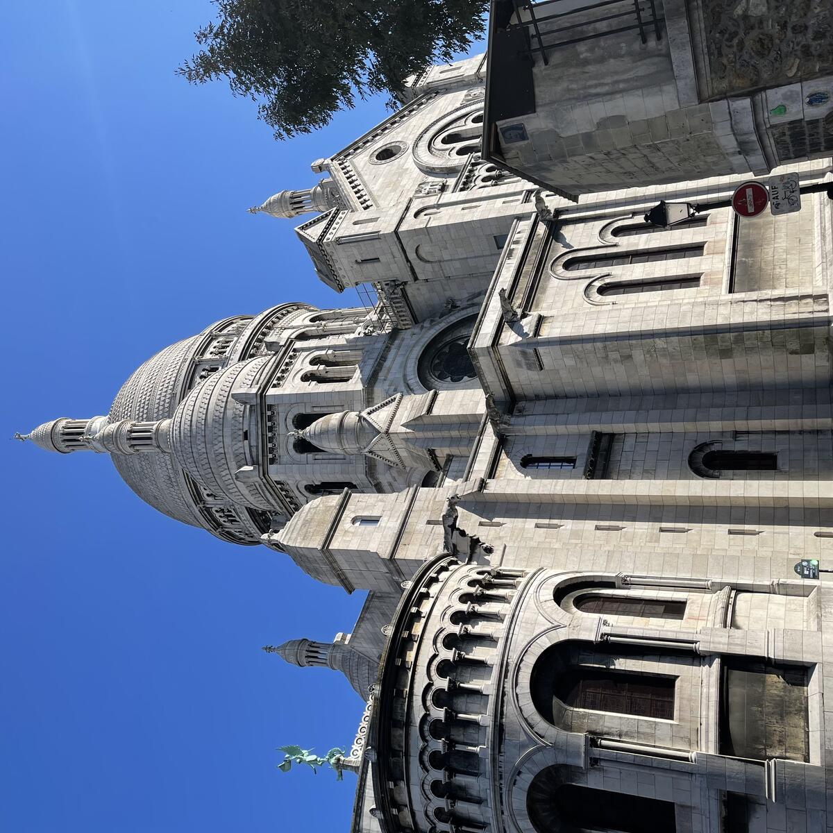 The large, white stone Sacré-Cœur Basilica in Paris stands under a clear blue sky, with its central dome and arcaded apse prominent, and people walking below.