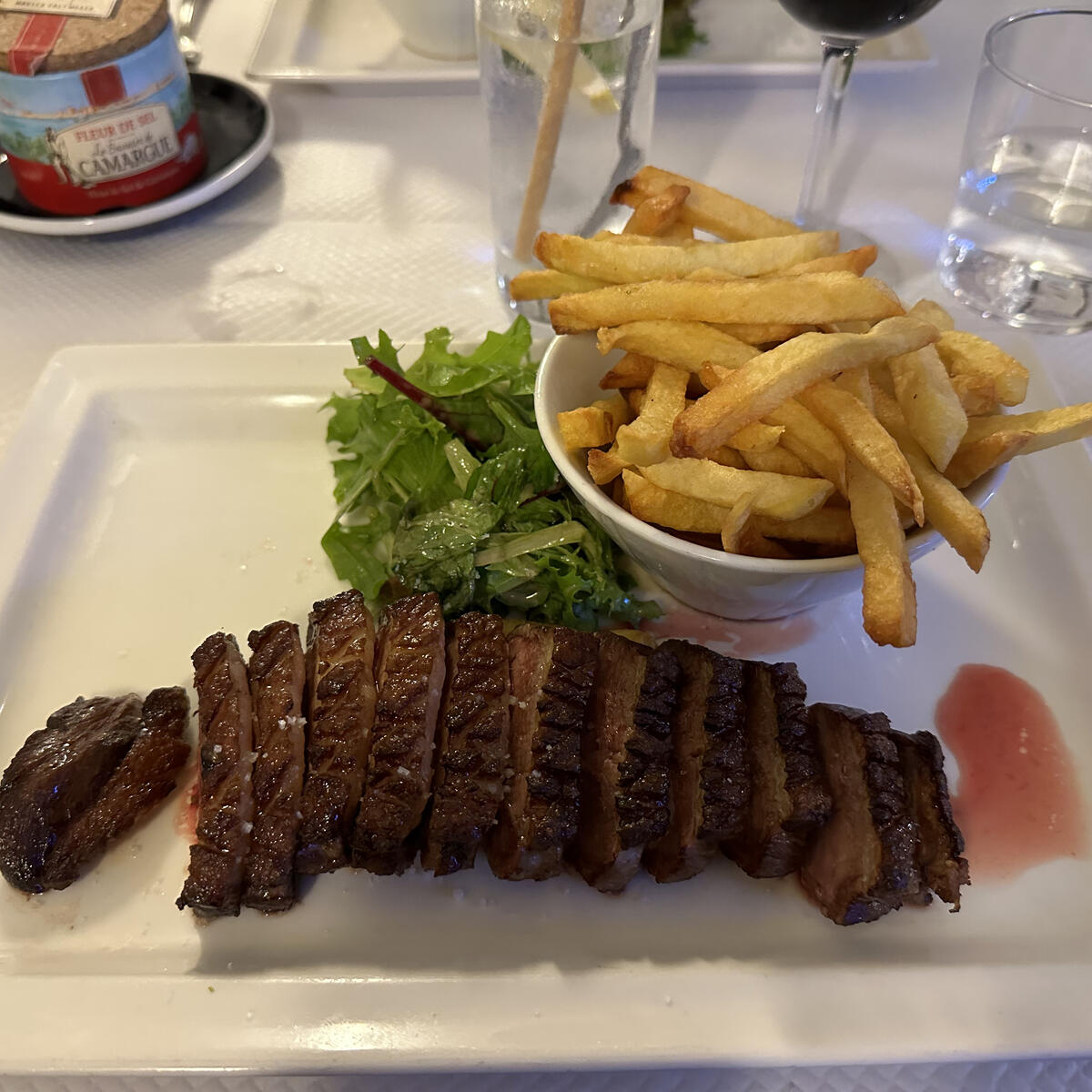 A white rectangular plate holds sliced grilled meat, a small bowl of golden french fries, and a side salad, set on a table with salt and water glasses.