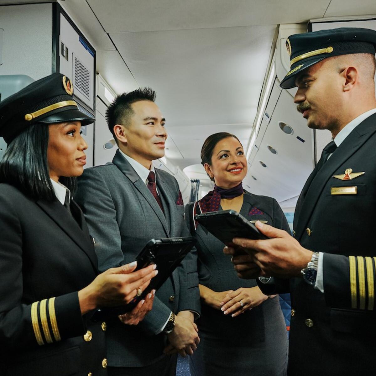 Two Delta pilots and two Delta flight attendants stand together in an airplane aisle, holding tablets and talking.