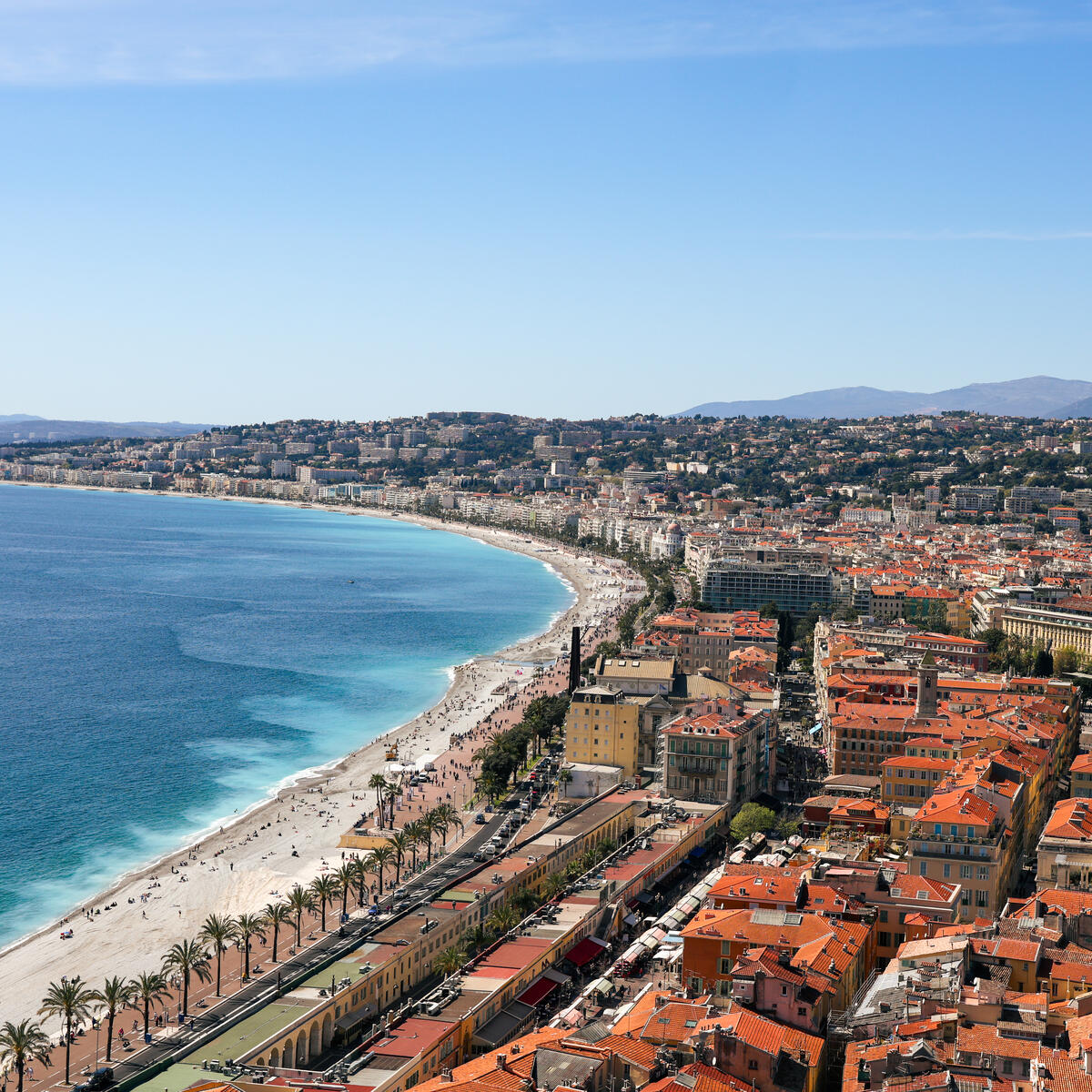 Aerial view of a curved Mediterranean beach and waterfront city with red-tiled roofs.
