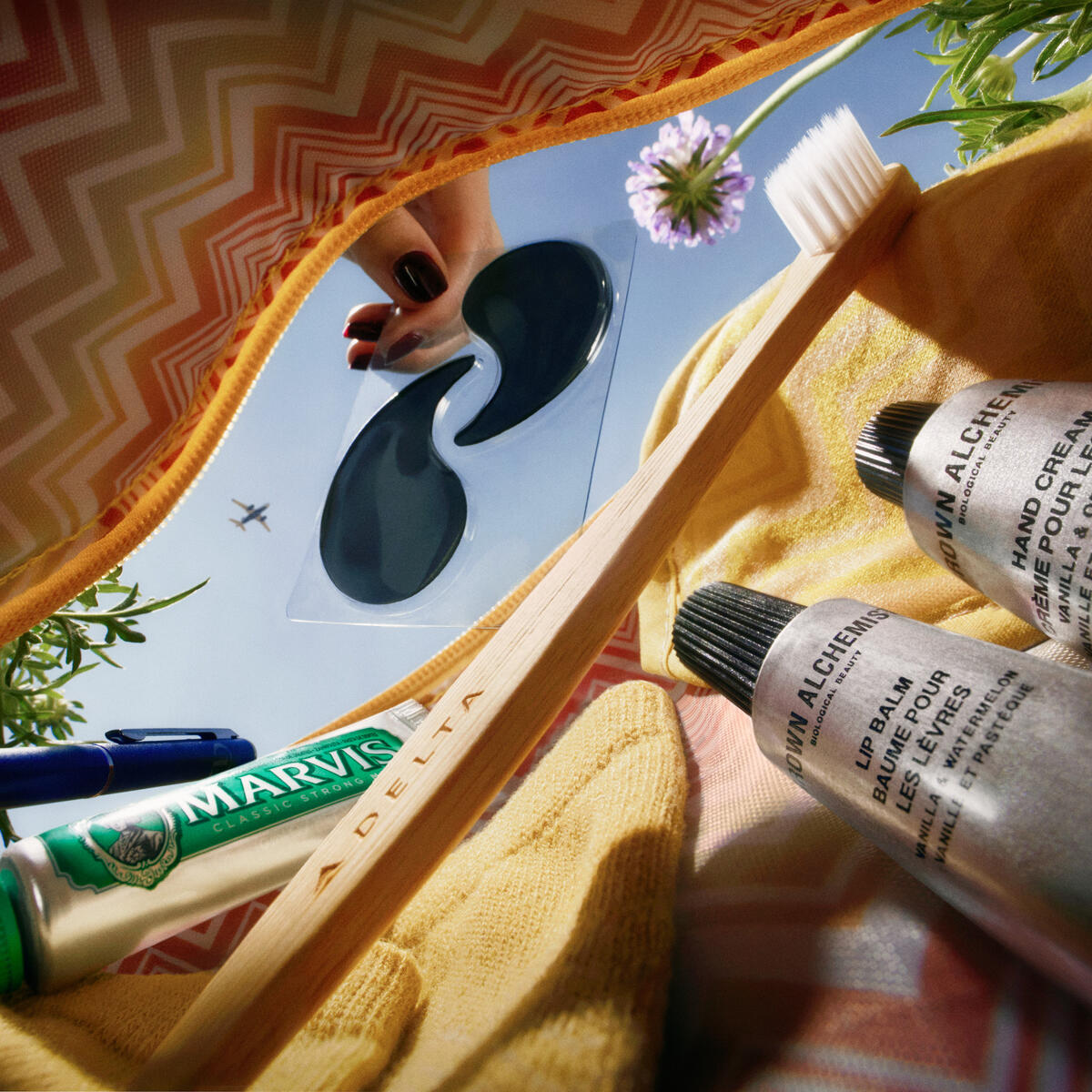 View inside a Missoni amenity kit showing a toothbrush, toothpaste, lip balm, hand cream, eye mask, and pen against a blue sky.