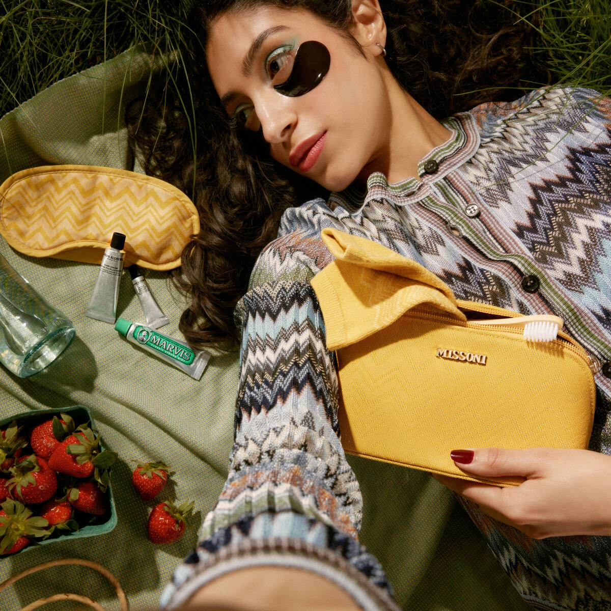 Person lying on grass with a yellow Missoni pouch, eye mask, skincare items, toothpaste, and strawberries arranged on a picnic cloth.