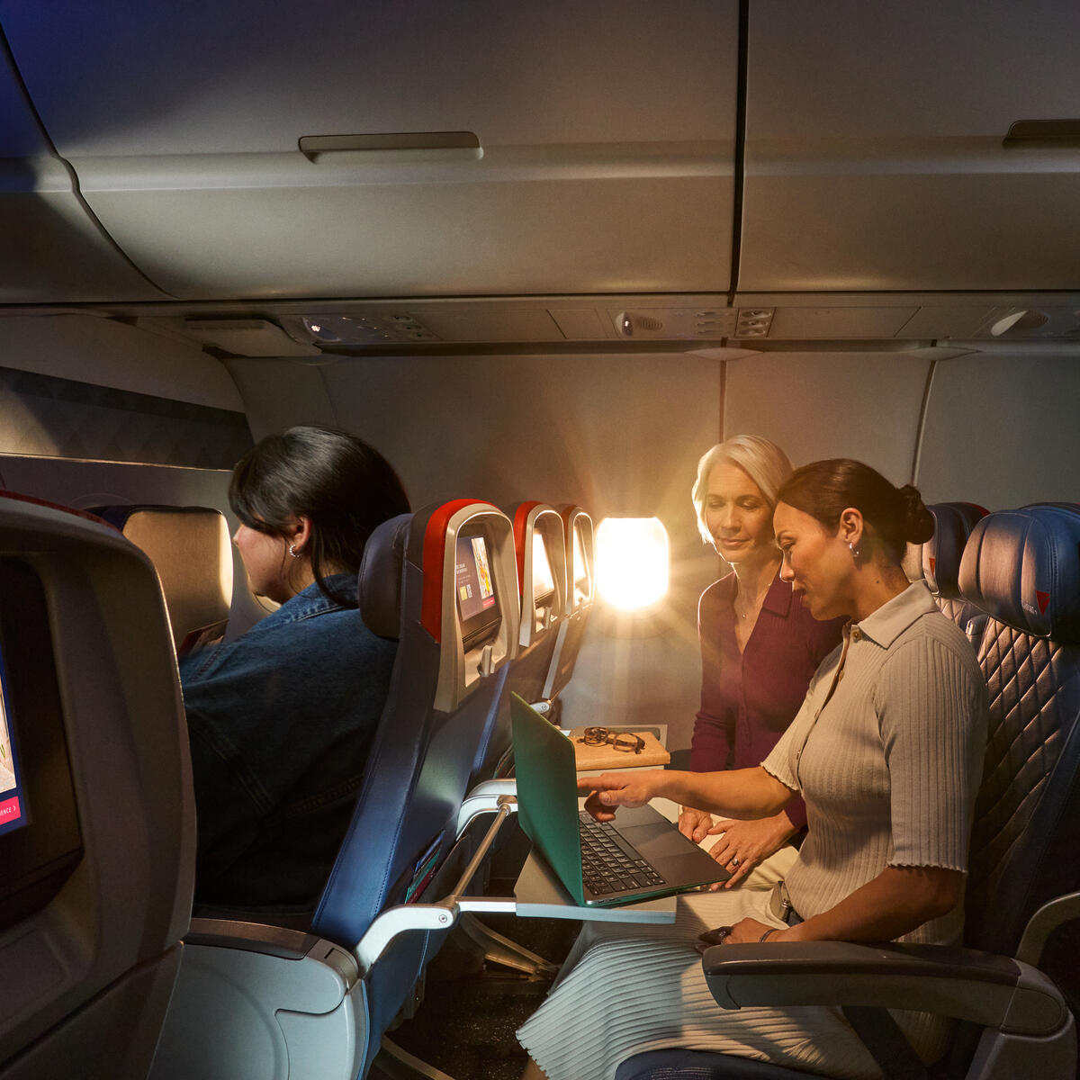 Two female Delta customers on board an aircraft working on laptops, with warm sunlight streaming through the windows.