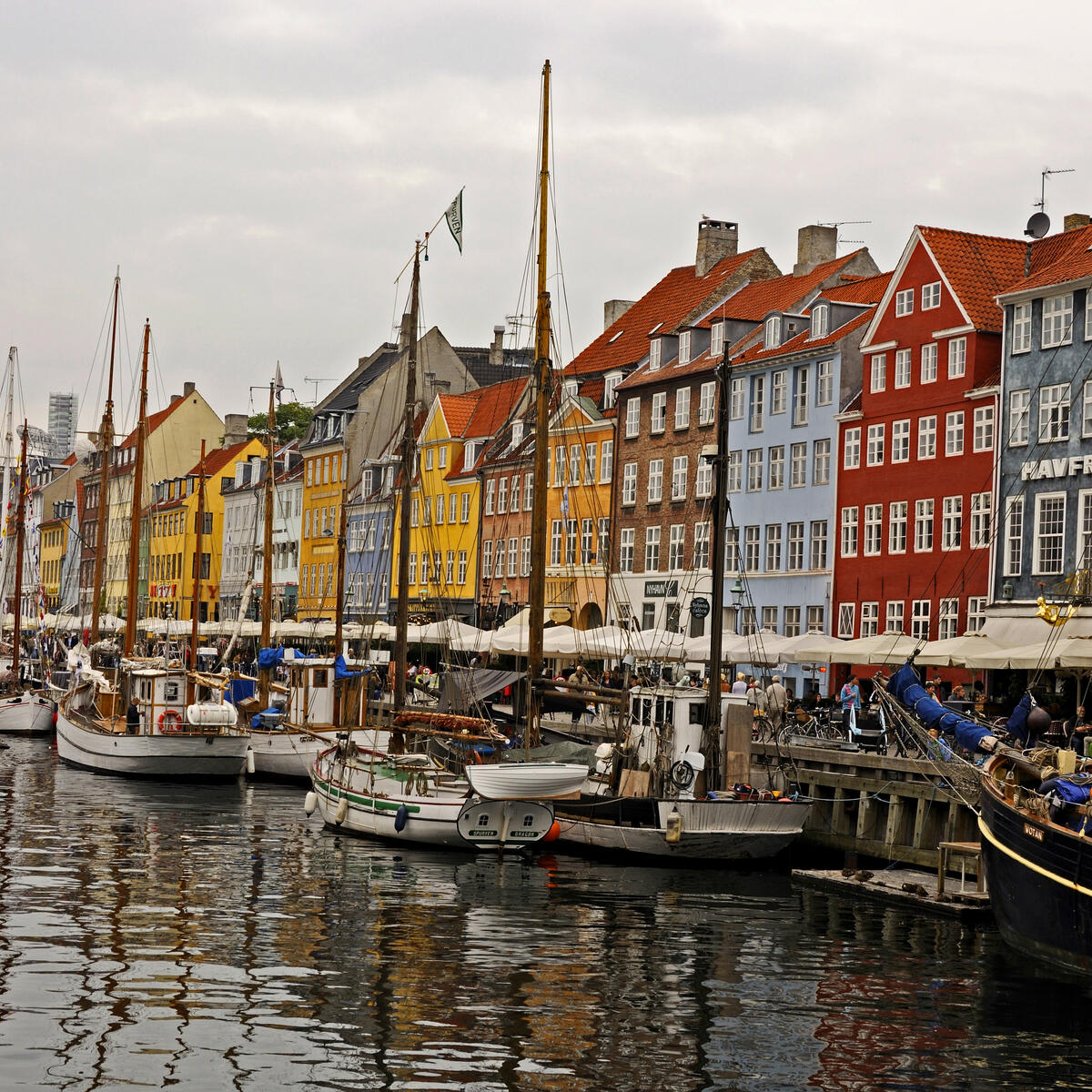 Colorful waterfront buildings line a canal with docked boats and masts, reflecting on the water under an overcast sky.