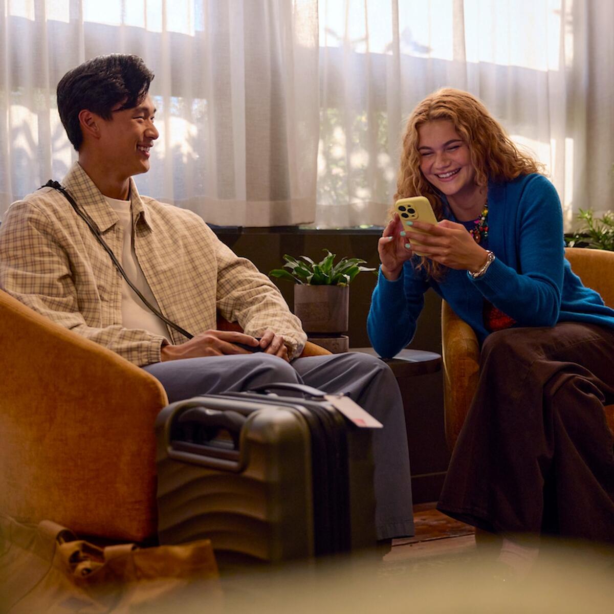 Two travelers sit in lounge chairs with rolling suitcases nearby, one looking at a smartphone, in a softly lit indoor waiting area with plants and sheer curtains.