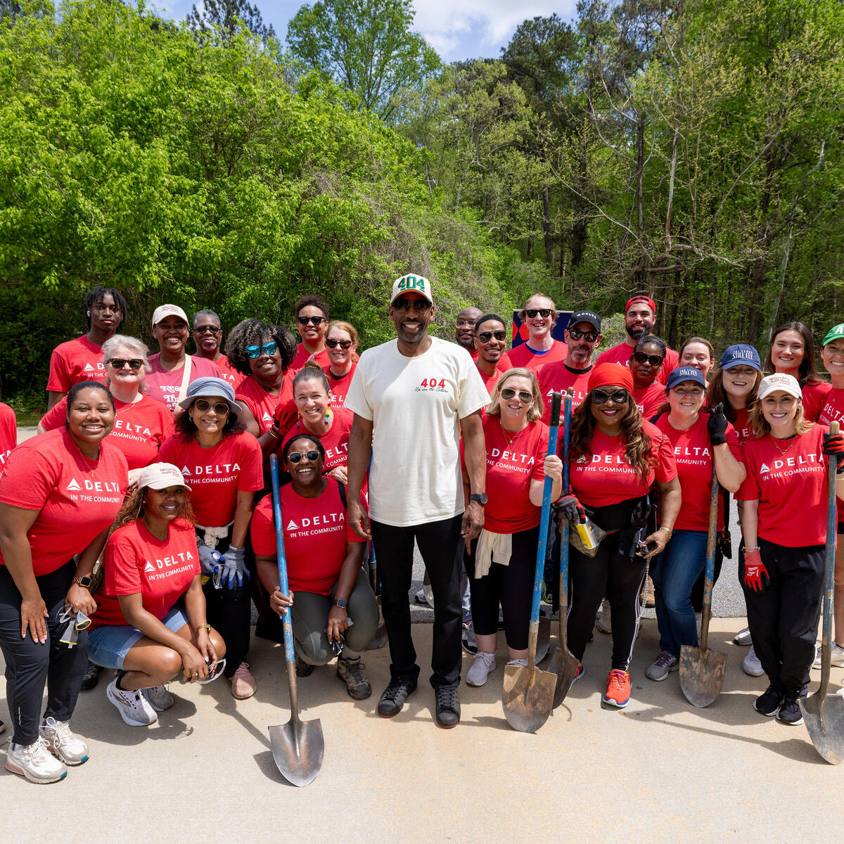 A large group of Delta volunteers wearing red “Delta in the Community” shirts stand together outdoors holding shovels, gathered on a paved area with trees in the background.